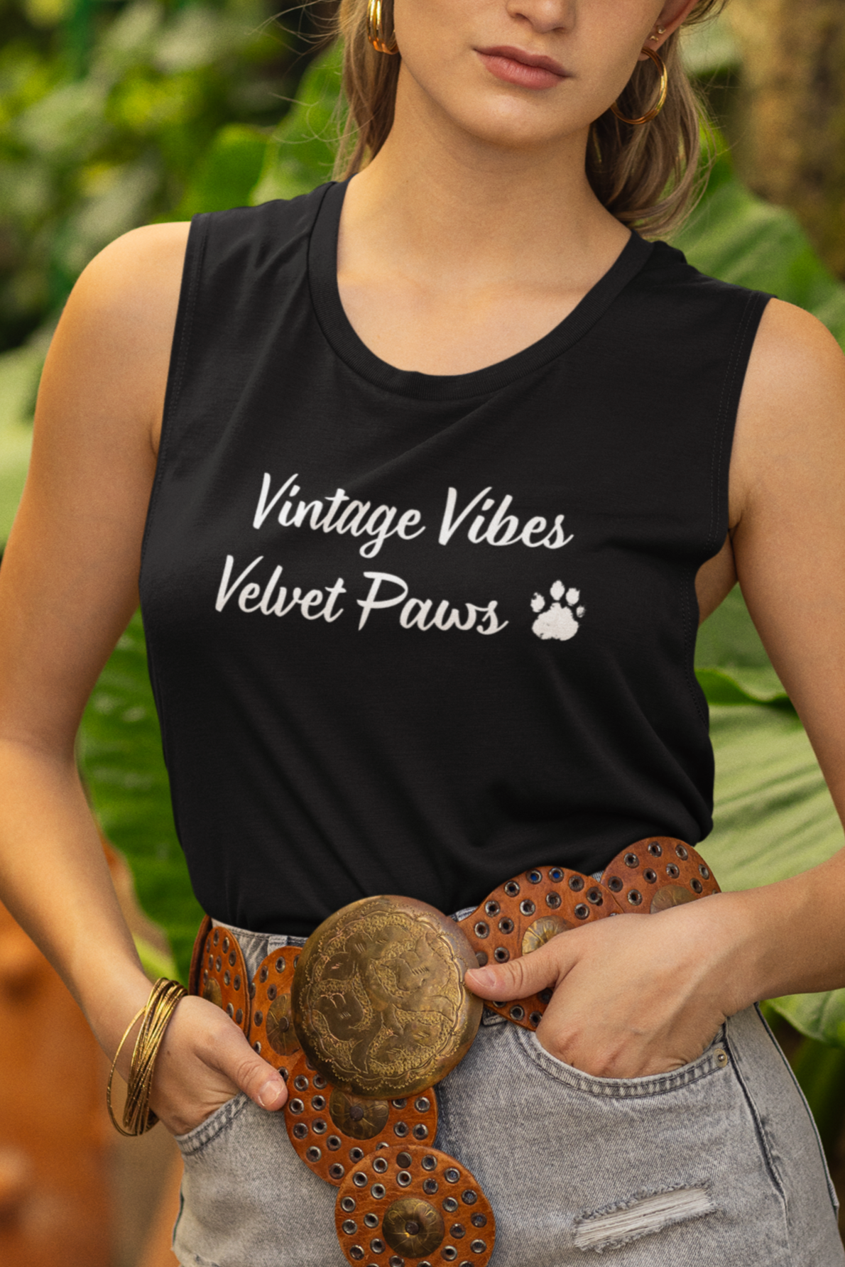 Woman wearing a black sleeveless shirt with text, denim shorts, and a brown hat in a garden setting.