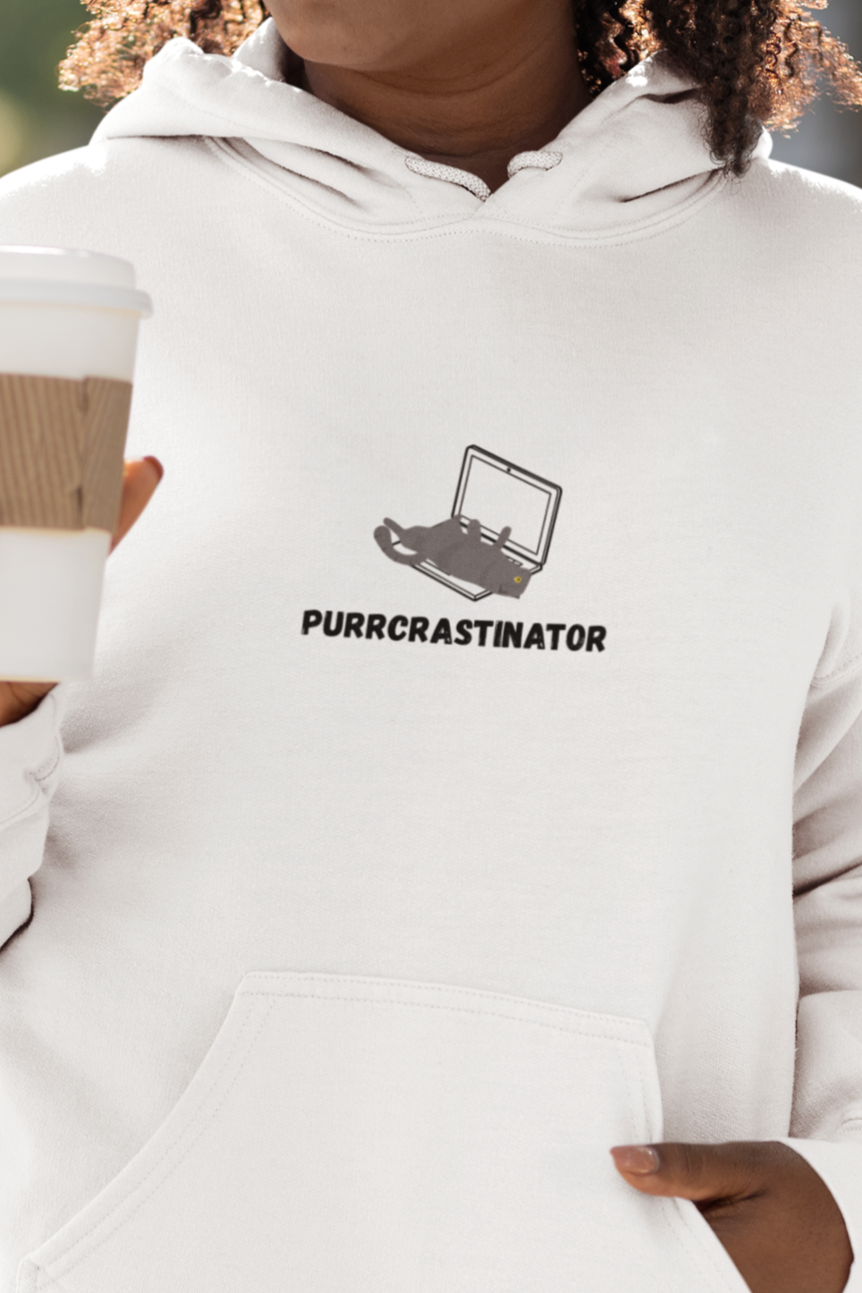 Woman wearing a white hoodie with a logo and text, holding a coffee cup outdoors.
