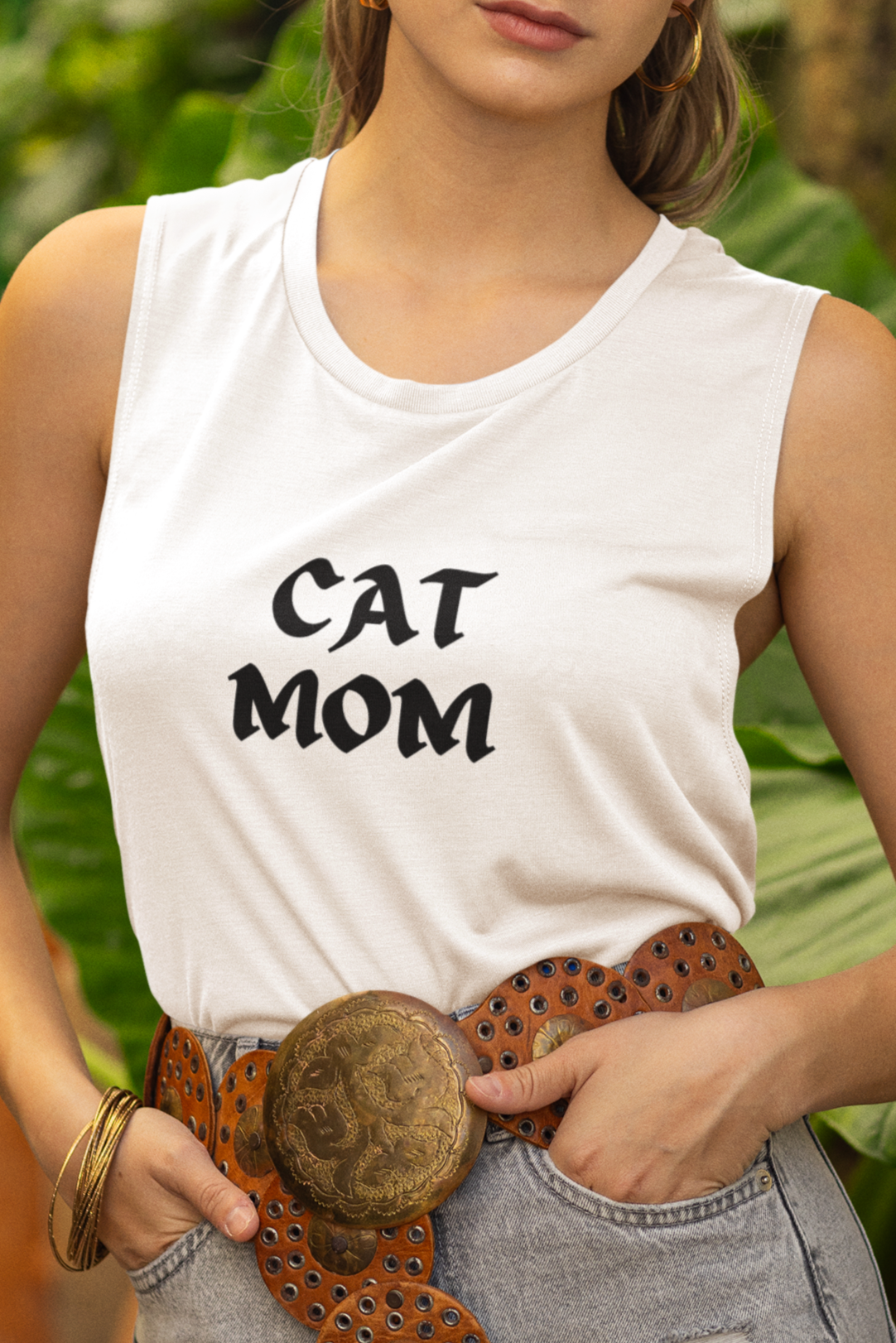 Woman wearing a white tank top with 'CAT MOM' text, standing outdoors with greenery in the background.