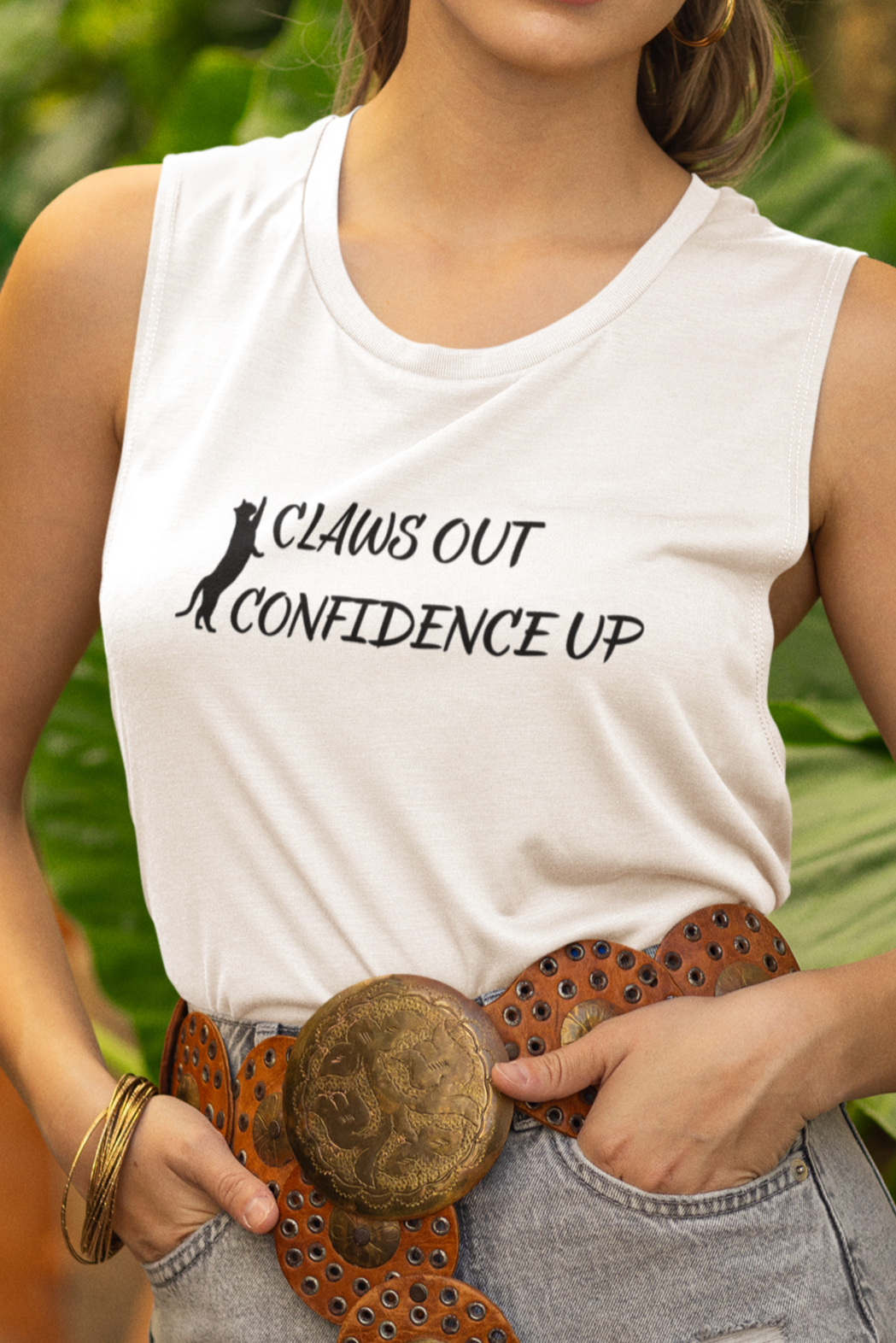 Woman wearing a white tank top with text, denim shorts, and a hat in a garden setting.