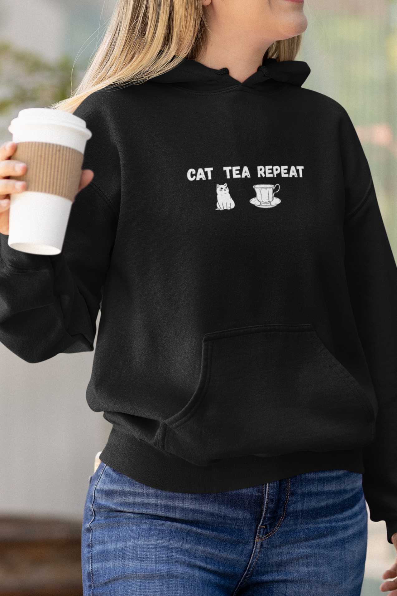 Woman wearing a black hoodie with text, holding a coffee cup outdoors.