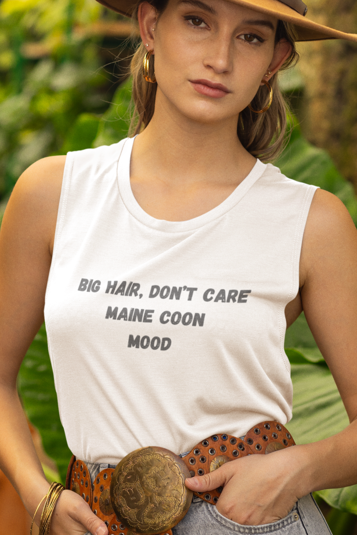 Woman wearing a white sleeveless shirt with text, denim shorts, and a brown hat in a garden setting.