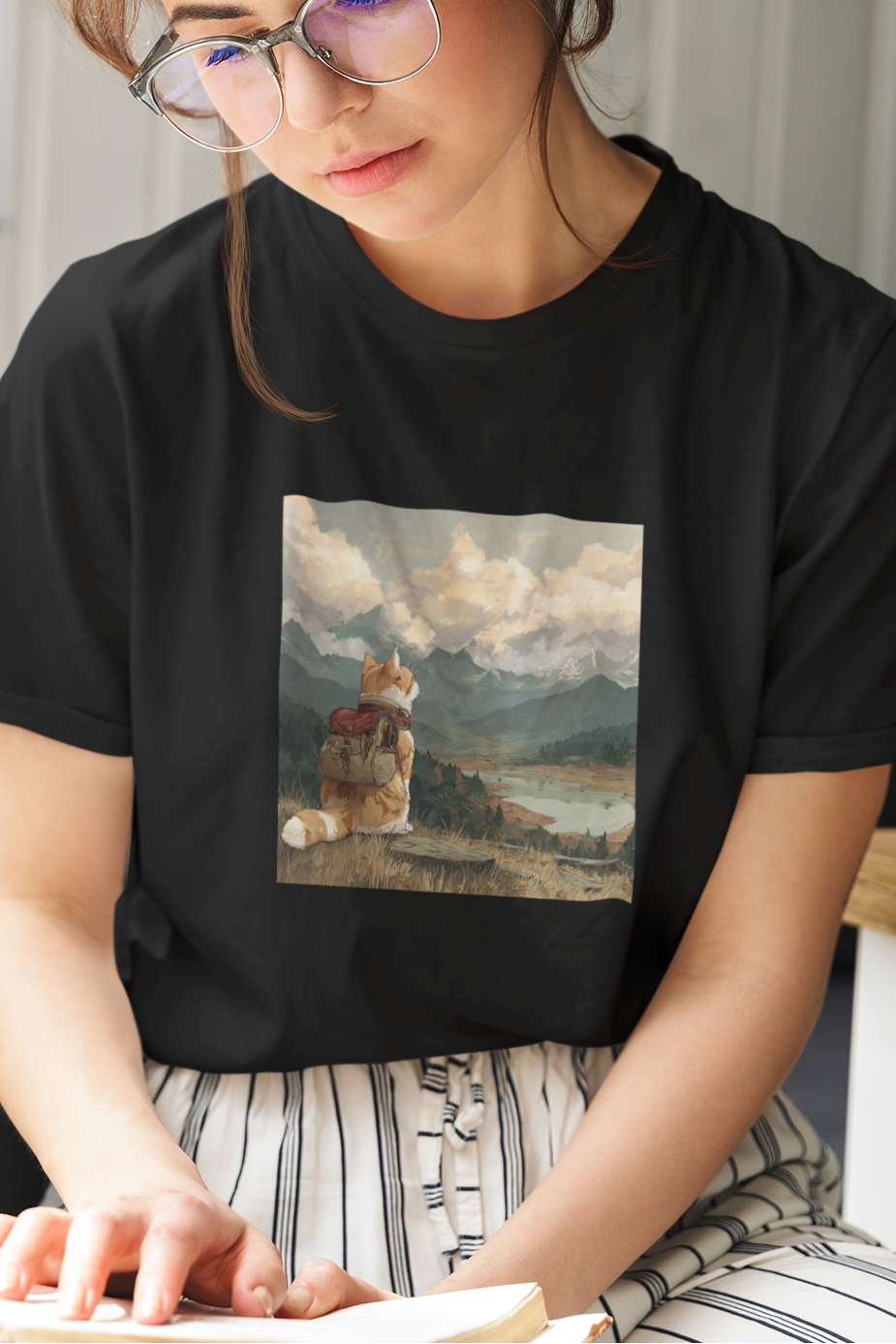 Woman reading a book while wearing a black t-shirt with a scenic design.